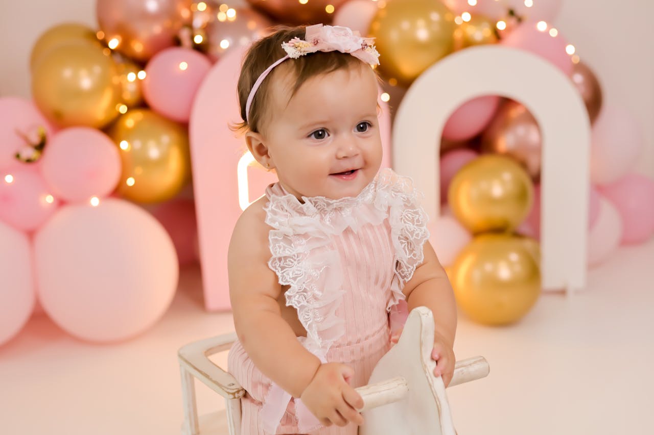 Charming baby girl smiling at a bright, festive birthday party with balloons.