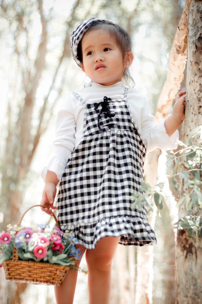 Adorable child in a black and white checkered dress holding a basket of flowers outdoors.